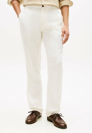 Mand iført hvide hørbukser og brune læder snørestøvler, med den ene hånd i lommen, stående på en hvid baggrund.