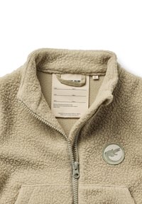 Veste en polaire beige avec un col haut, fermeture éclair et un logo circulaire vert. Présente un matériau texturé et une étiquette d'entretien à l'intérieur.