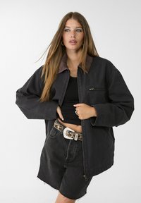 Zwarte oversized jas met een ribbelkraag, rits sluiting, en details van voorzakken, gedragen over een zwart top en hoge zwarte denim shorts.