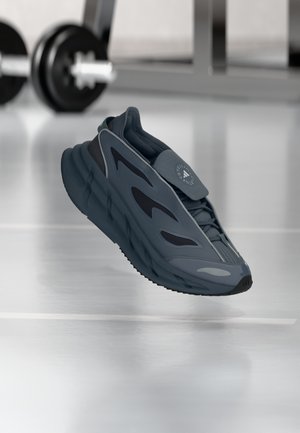 Gray athletic shoe featuring a textured upper with mesh accents, rounded sole, and a logo detail on the tongue, set against a gym background.