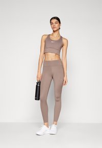 Smeđi sportski crop top i tajice viskog struka, glatka tekstura, Nike logo na oba komada, u pratnji crne bočice za vodu i bijelih tenisica.