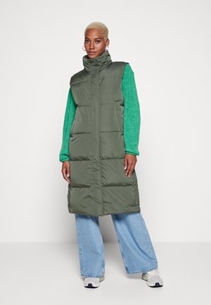 Gilet rembourré vert avec col montant, fermeture éclair sur le devant et poches latérales, porté sur un pull vert et un jean bleu large. Détail de tissu texturé visible.
