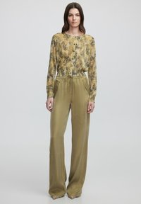 Pantalones largos de color verde oliva, con un acabado satinado, bolsillos laterales y cintura con cordón, combinados con una blusa de botones amarilla y verde con estampado.