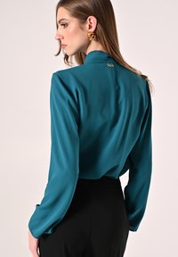 Blusa di raso teal con maniche lunghe e colletto alto, caratterizzata da un logo discreto sul retro e una texture liscia e lucida.