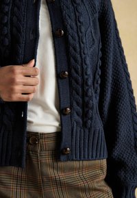 Navy adīts džemperis ar kabeli un tekstūras rakstiem, ar brūnām pogām un ribotu apakšmalu, valkāts pāri baltam topam un raibām biksēm.