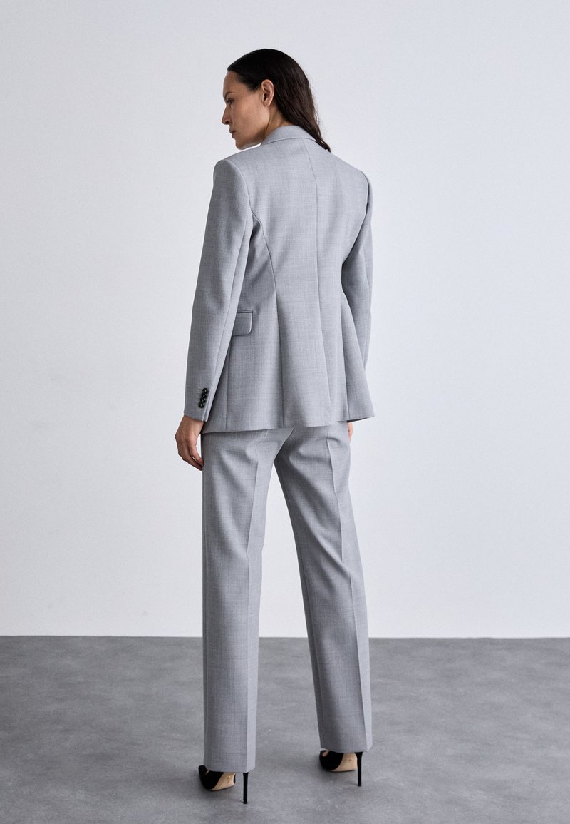 Szary garnitur skrojony na miarę z usztywnianą marynarką i prostymi spodniami. Tkanina ma gładką teksturę, z delikatnym wzorem kraty i eleganckimi czarnymi szpilkami.