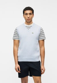 Άνδρας που φορά ανοιχτό μπλε γιλέκο πλεχτό με καλώδια πάνω από μια μαύρο-λευκή ριγέ κοντομάνικη μπλούζα και μαύρο σορτς, στεκόμενος μπροστά σε απλό λευκό φόντο.