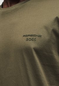 Κοντινό πλάνο σε μπλουζάκι ελιάς με ανάγλυφο λογότυπο κειμένου "PORSCHE BOSS" στο στήθος.