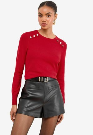 Pull en tricot rouge à manches longues, avec des boutons décoratifs blancs à l'épaule. Associé à un short en cuir noir taille haute.