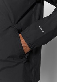 Manche de veste noire avec le logo "WINDWALL", main placée à l'intérieur d'une poche zippée sur le côté, montrant un tissu noir et un poignet élastique.