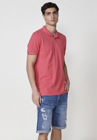 Polo majica s kratkimi rokavi v svetli koralni barvi z dvema gumboma, v kombinaciji z raztrganimi denim kratkimi hlačami z zavihanimi robovi in vozlanjem tkanine.