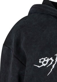 Juodas džemperis su unikaliu tekstūruotu audiniu. Priekyje yra didelė, nusidėvėjusi balta grafika. Matoma kapučino konstrukcija.