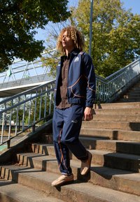 Mornarska trenirka z oranžnimi detajli, ki vključuje zip jakno in usklajene hlače. Obleka je kombinirana s bež športnimi čevlji, na zunanjih stopnicah.