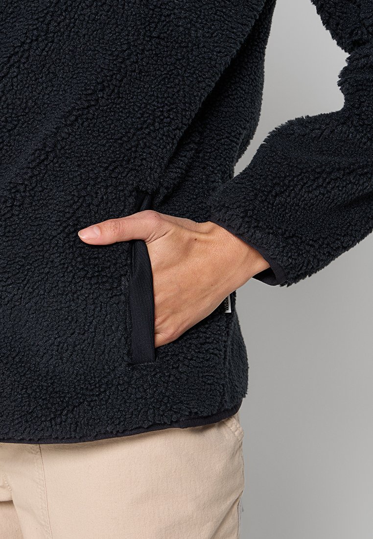 Main dans une poche de veste en polaire texturée noire, pantalons beiges visibles, fond neutre.