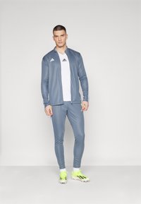 Gray athletic jacket with white stripes, paired with a white T-shirt and gray pants. Features neon green soccer cleats with black accents.