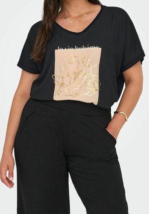 Femme portant un t-shirt noir à manches courtes et col en V avec un motif floral doré et du texte, associé à un pantalon noir et un bracelet doré.