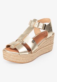 Sandalia cuña metálica dorada con plataforma de yute trenzado, presenta tiras entrecruzadas y cierre de hebilla, textura suave similar al cuero.