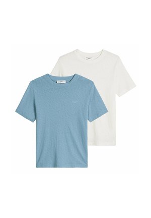 Hombre que lleva una camiseta blanca lisa de manga corta, vaqueros azul claro, collar y pulsera de cadena plateada, con una mano en el bolsillo.