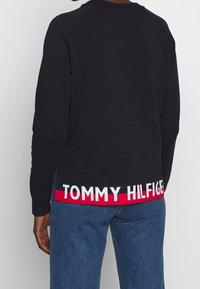 Felpa nera con collo rotondo e maniche raglan, caratterizzata da una banda con logo rosso e bianco sul bordo. Indossata con jeans blu.