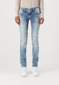 Femme portant un jean skinny bleu clair délavé avec une taille à double bouton et des baskets blanches, debout devant un fond uni.