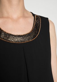 Svart ärmlös topp med en bred, metallisk svart och guld texturerad krage med intrikata detaljer och en subtil plissé nedanför halsringningen.