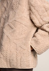 Pulover iz kremastega flisa z teksturirano, vzorčasto površino in nevtralnim robom. Ima ovratnik z zadrgo do polovice in stranske žepe.