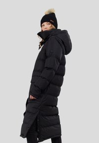 Schwarze Puffermantel mit langem, gepolstertem Design, ausgestattet mit einer Kapuze und seitlichen Schlitzen. Begleitet von einer schwarzen Strickmütze mit einem Bommel.