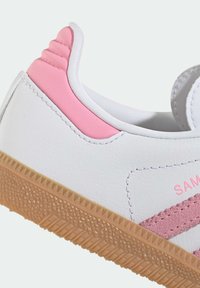 Biele kožené tenisky s ružovými akcentmi, s textúrovanou podrážkou v zlatej farbe a diagonálnymi pruhmi. Hladký povrch so šitými detailmi.