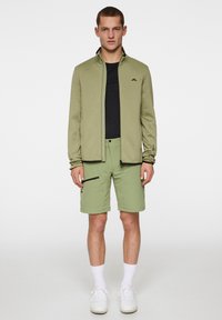 Oliwkowa kurtka z zamkiem błyskawicznym i czarnymi akcentami, zestawiona z dopasowanymi szortami z bocznymi zamkami i luźnym krojem. Model nosi białe trampki.