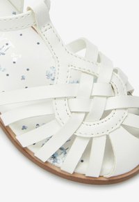 Sandale en cuir blanc avec des lanières entrelacées, une silhouette à bout rond et une semelle intérieure à motif floral avec des accents bleus.