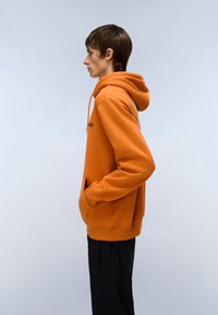 Felpa arancione realizzata in un tessuto morbido, con tasca a marsupio, polsini a coste e cappuccio con coulisse. Indossata con pantaloni neri. Vista di profilo.