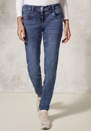 Jeans skinny fit en denim bleu foncé, avec une fermeture boutonnée, cinq poches et des détails de couture minimalistes. Portés avec des baskets beiges.