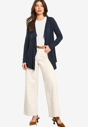 Cardigan aperto navy con maniche lunghe, frontale drappeggiato, abbinato a una camicia bianca e pantaloni a gamba larga color crema, con cintura e tacchi marroni.