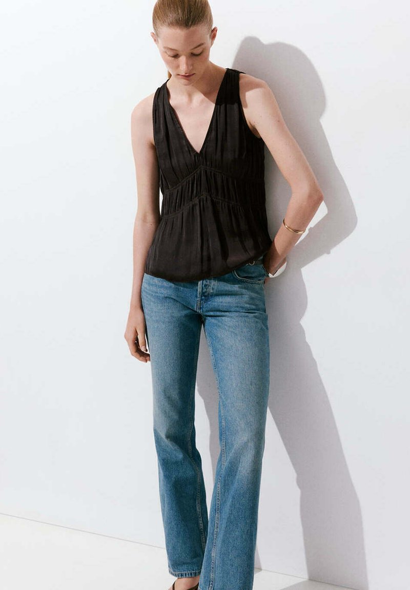 Femme portant un haut noir sans manches à encolure en V et un jean bleu taille haute, debout contre un mur blanc avec une main dans la poche.