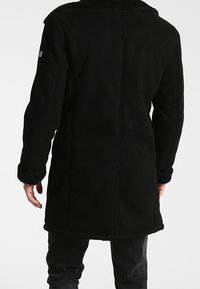 Manteau en polaire noire avec une finition texturée, doté d'un large col, de manches longues et d'une doublure en fourrure aux poignets et au col. Vue de dos.