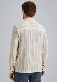 Chemise beige clair avec des rayures texturées verticales, poignets à boutons et col. Le tissu semble doux et léger, idéal pour le superposition.