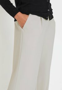 Weite Hosen in hellem Beige, aus glattem Stoff gefertigt, mit seitlichen Taschen. Kombiniert mit einer schwarzen Strickjacke, die Knopfdetails zeigt.
