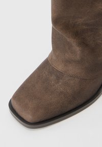 Brown suede knee-high boot with a square toe, subtle texture, and a black rubber sole. Seam detail along the upper  enhances the design.