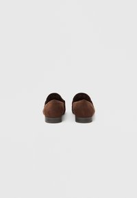 Mocassins en daim marron avec un petit talon et un bout arrondi, présentant une texture lisse et des détails de couture minimaux à l'arrière.
