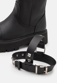 Bottine en cuir noir avec une semelle épaisse, associée à un harnais en cuir noir agrémenté de clous argentés et de boucles métalliques réglables.