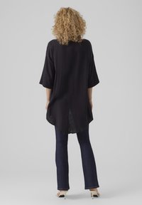Tunic nera e dal texture particolare con maniche a tre quarti, caratterizzata da un orlo posteriore più lungo. Abbinata a jeans scuri e scarpe con tacco di colore chiaro.