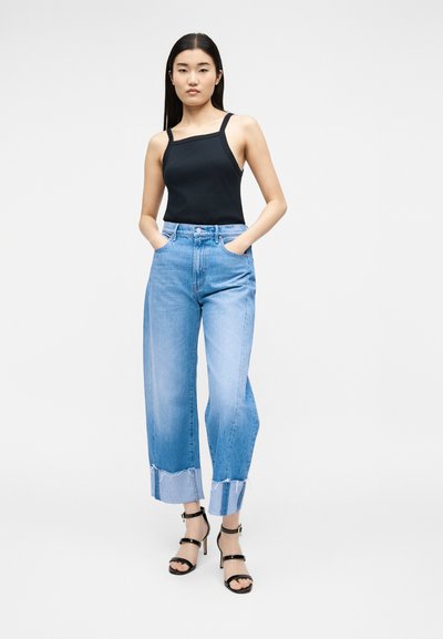 Νεαρή γυναίκα που φοράει μαύρο αμάνικο τοπ, ανοιχτό μπλε φαρδιά cropped τζιν με ατημέλητες άκρες και μαύρα σανδάλια με ψηλό τακούνι, στέκεται με τα χέρια στις τσέπες.