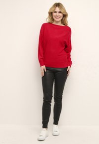 Maglione rosso a maniche lunghe con scollo a barca, abbinato a pantaloni neri aderenti e sneakers bianche. Tessuto morbido; design informale.