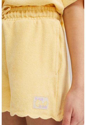 Bløde gule terry-cloth shorts med elastisk talje, bølget kant, forlomme og et mærke med teksten "Le Club De Soleil" samt en citron-grafik.