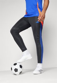 Schwarze Sporthosen mit blauen Seitenstreifen, kombiniert mit einem blauen Oberteil und weißen Fußballschuhen, stehend auf einem schwarz-weißen Fußball.