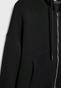 Sweat à capuche zippé noir avec poche kangourou à l’avant, capuche avec cordon de serrage et fermeture éclair argentée visible sur le côté droit du vêtement.