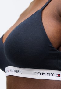 Čierny bavlnený bralette s tenkými ramienkami a bielym elastickým pásom s nápisom "TOMMY HILFIGER" a detailom loga. Hladká textúra.