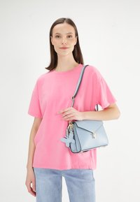 Jasnoniebieska torebka na zamek z klapą, z złotymi akcesoriami i breloczkiem w kształcie motyla, połączona z luźnym różowym t-shirtem i dżinsami.