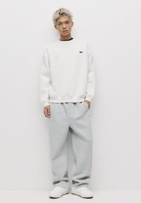 Sweatshirt blanc à col rond sur une chemise noire, associé à un pantalon de survêtement gris clair et des baskets blanches. Textures simples, branding minimal.