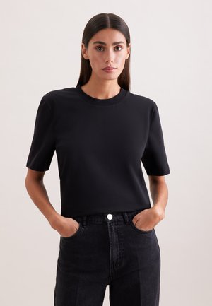 Femme aux cheveux bruns lisses, portant un t-shirt noir uni à manches courtes et un jean taille haute foncé, les mains dans les poches, fond neutre.
